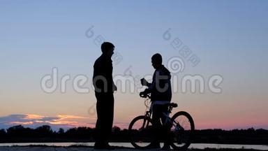 那家伙拿起电话，在黎明时在电话上自拍。 一个男子和一个骑自行车的少年的剪影